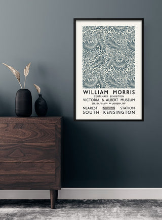 Framed William Morris grey poster with botanical design hanging on a dark wall above a wooden dresser.