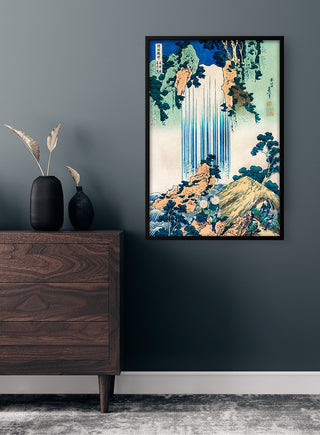 Framed "Yoro Waterfall" by Katsushika Hokusai hanging on a dark wall above a wooden dresser.