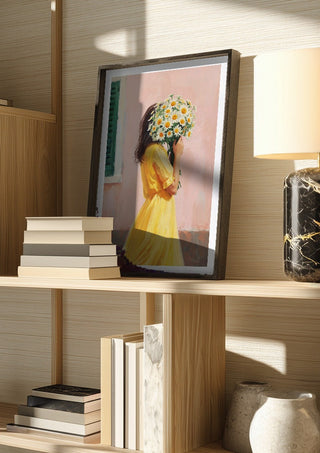 Girl in yellow dress with flowers by Ohkimiko in a dark frame on a wooden shelf beside books and a lamp