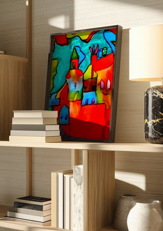 The Village by Nittaart in a dark frame on a wooden shelf beside books and a lamp