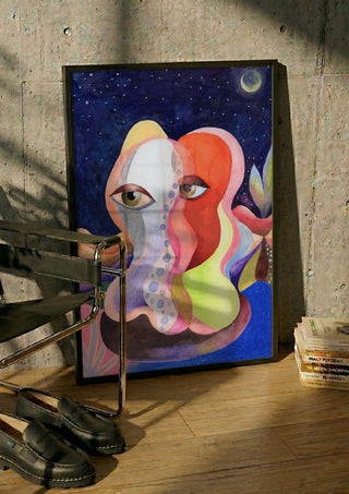 Two Faces Under the Moon by Shimmering Ugly in a black frame leaning against a concrete wall by a steel chair