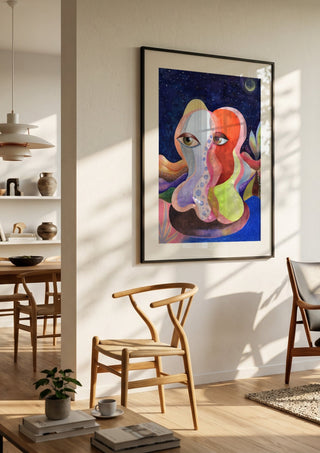 Two Faces Under the Moon by Shimmering Ugly in a black frame in a bright dining room with wooden chairs
