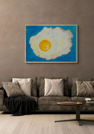 Fried egg by Sulaika Martis in a light oak frame on a warm grey wall above a brown sofa