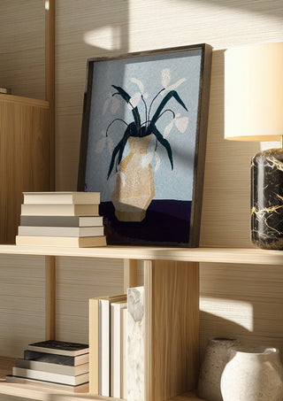 Vita blomster i beige vas by Rebecka Hansson in a dark frame on a wooden shelf beside books and a lamp