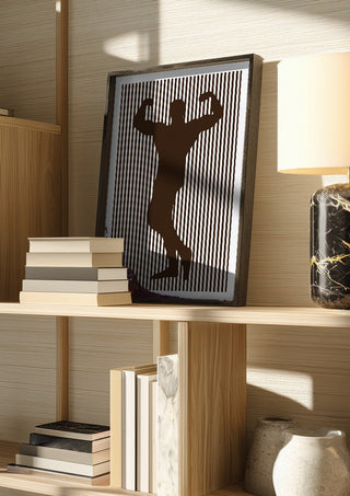 bodybuilder silhouette by nyai in a dark frame on a wooden shelf beside books and a lamp