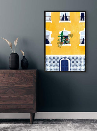 Framed artwork of a yellow Lisbon building by Maja Tomljanovic displayed on a dark wall above a wooden dresser.