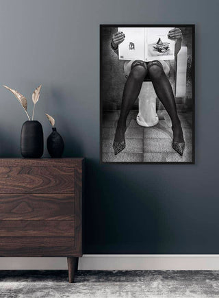 Framed print of a person in stockings sitting on a toilet reading, displayed on a modern dark wall.