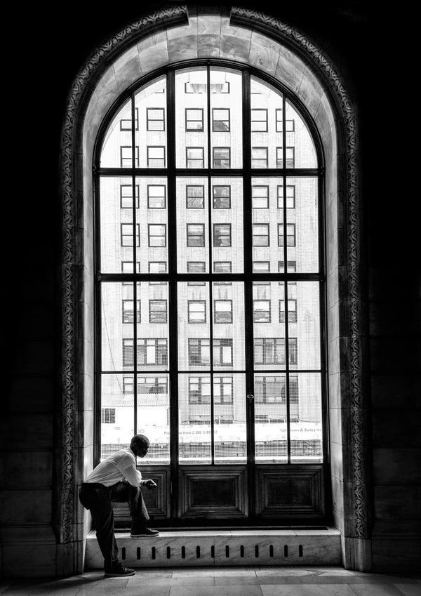 A man stands alone by a tall arched window, gazing at city buildings outside.