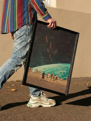 Person holding framed "The Others" artwork by Frank Moth, featuring a surreal beach and space scene.