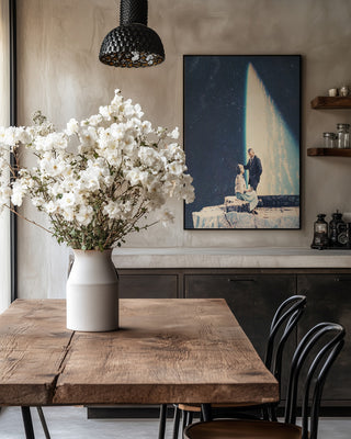 Framed "This Love" by Frank Moth hanging in a cozy dining room with a wooden table and white flower centerpiece.