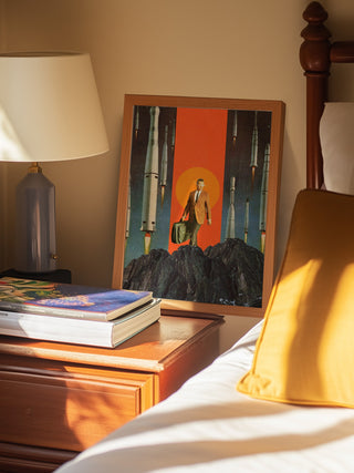 Framed print of "The Departure" by Frank Moth leaning on a bedside table next to books and a lamp.