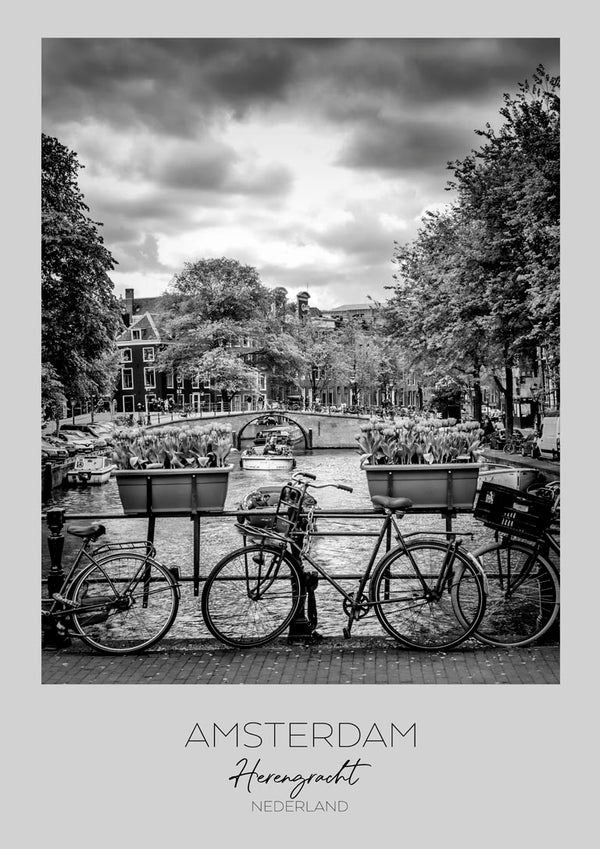 Black and white photo of bicycles by a canal on Herengracht, Amsterdam, with trees and buildings in the background.
