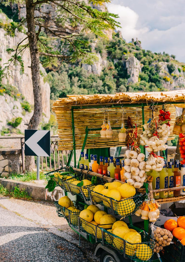 Street-side stall with lemons, garlic, and beverages on Amalfi Coast Drive with scenic cliffs in the background.