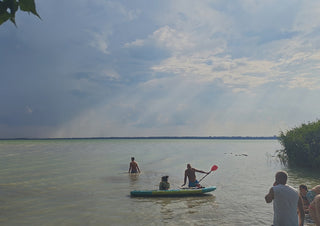 The Lake’s Shared Rhythm by Bora Kőhalmi - swimmers and a paddleboarder on the green water of Lake Balaton under a wide cloudy sky