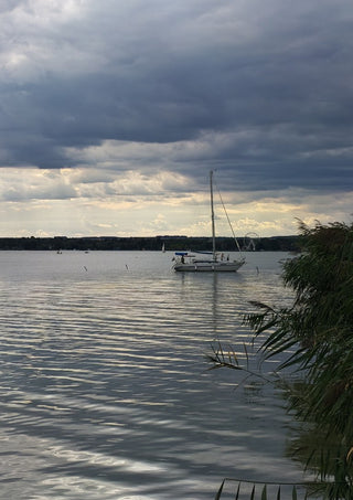 Light Reaching the Water by Bora Kőhalmi - a white sailboat under a heavy grey cloud with a band of light over Lake Balaton