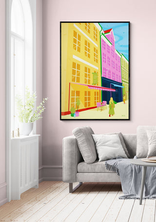 Framed "Under Blue Sky - Nanna Paavig" artwork with colorful buildings displayed above a modern sofa in a bright room.
