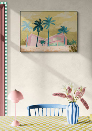 Framed artwork of The Pink House by Goed Blauw hanging above a table with flowers and a pink lamp.
