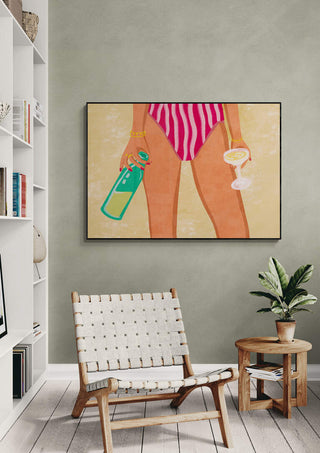 Framed art of a woman in stripes holding wine and bottle at the beach by Raissa Oltmanns, on display above a chair.