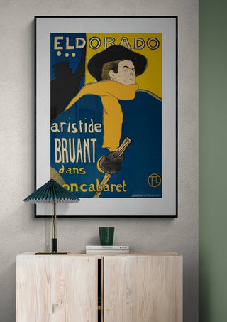 Framed Aristide Bruant cabaret poster by Lautrec displayed above a light wooden cabinet with lamp and cup.