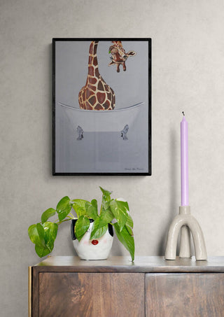Framed artwork of a giraffe in a bathtub by Coco de Paris, displayed above a cabinet with decor items.